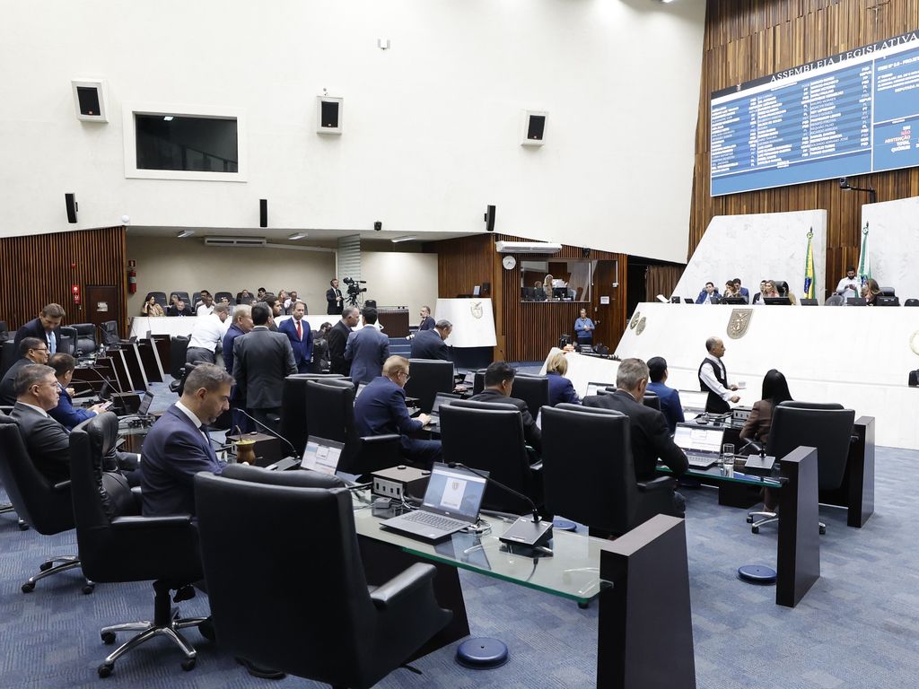 Plenário da Assembleia Legislativa do Paraná durante sessão, com deputados nas bancadas de cadeiras pretas, notebooks sobre as mesas e o painel de votação ao fundo
