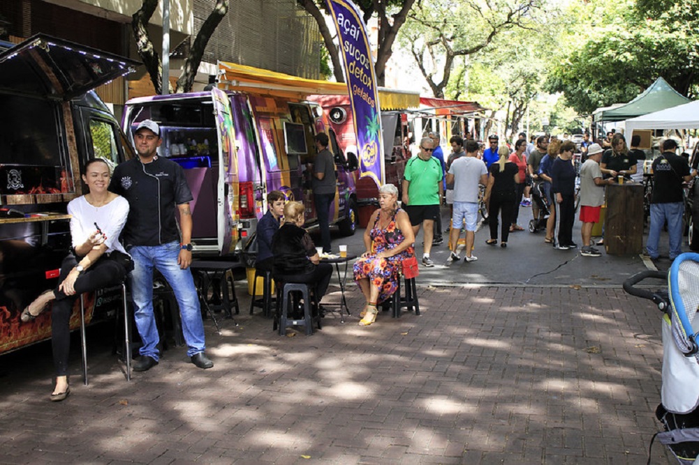 Food trucks coloridos estacionados em uma rua arborizada de Belo Horizonte, com pessoas sentadas em banquinhos e outras circulando entre os veículos e as tendas