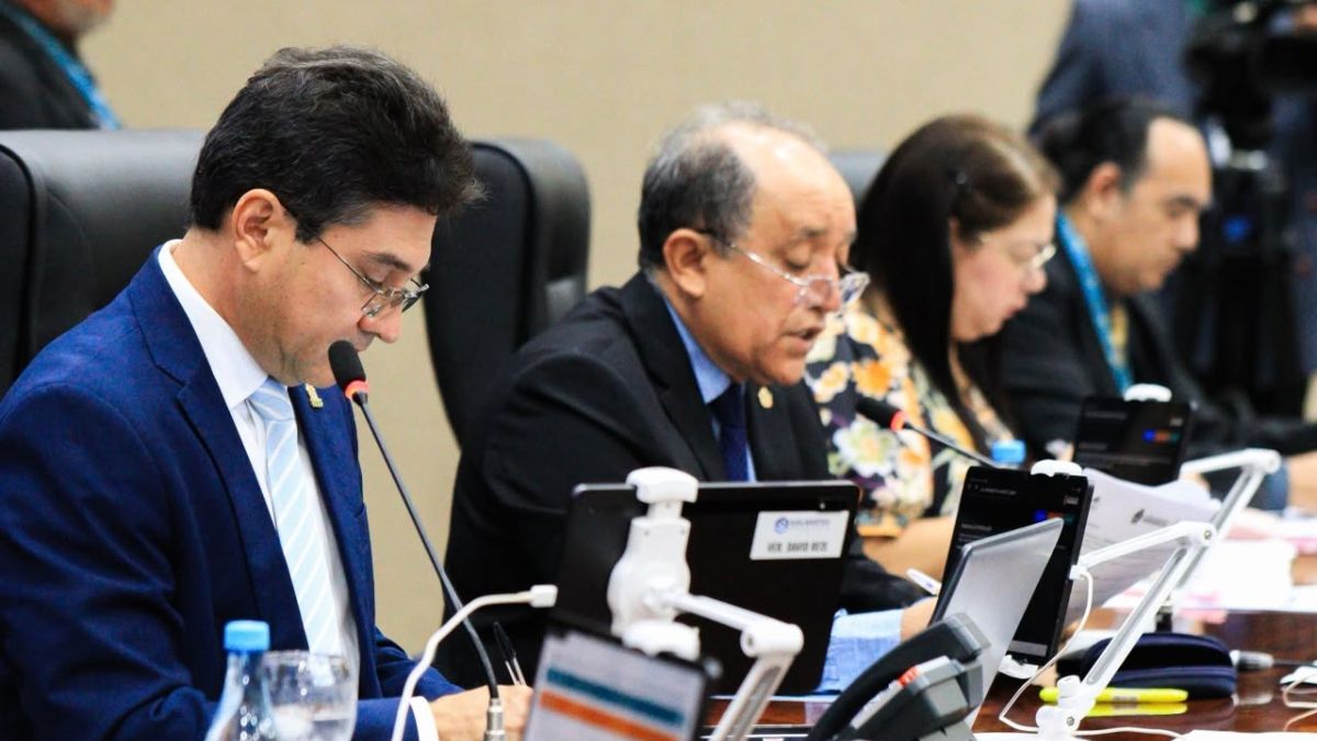 Vereadores sentados à mesa do plenário da Câmara Municipal de Manaus leem documentos diante de microfones durante sessão