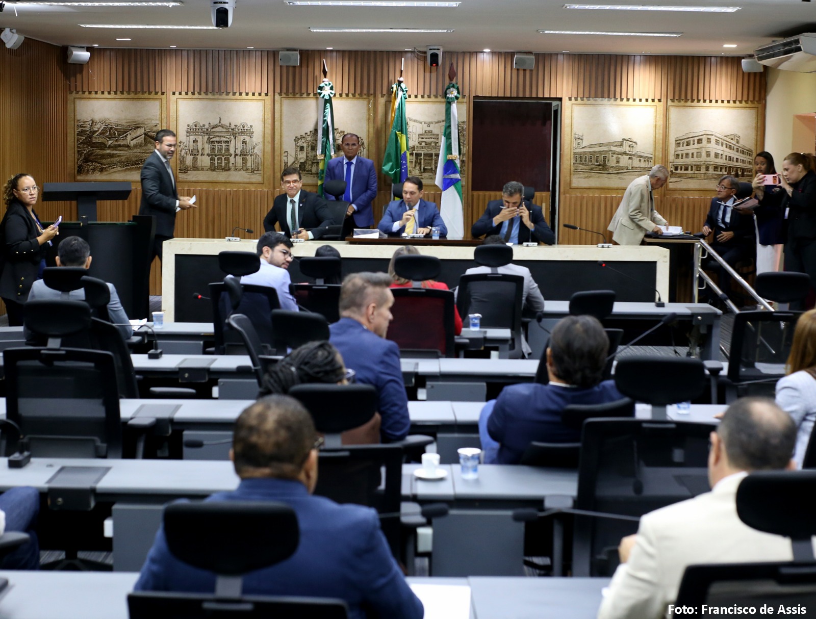 Plenário da Câmara Municipal de Natal durante sessão ordinária, com vereadores de costas nas bancadas e a Mesa Diretora ao fundo, diante de bandeiras, entre elas a do Brasil