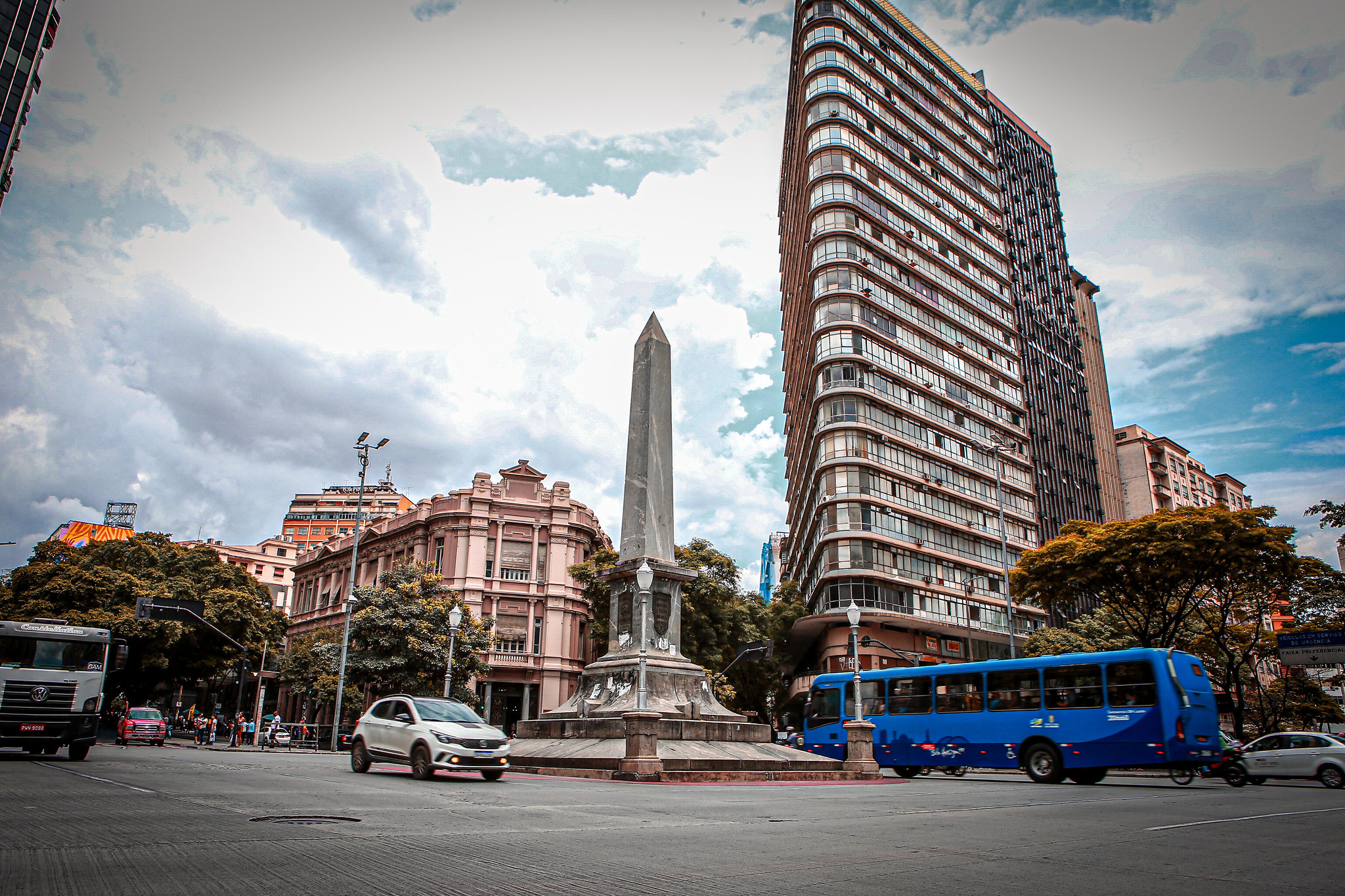 Praça Sete de Setembro, no Centro de Belo Horizonte, com o obelisco ao centro, prédios antigos e altos ao redor, carros e um ônibus azul circulando